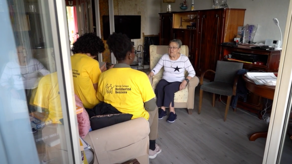 deux jeunes personnes avec un tshirt jaune discutent avec une vieille dame assis dans un canapé.